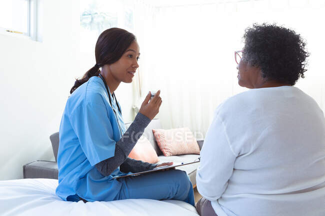 Старша змішана жінка проводить час вдома, відвідуючи змішану расу медсестру, сидячи на ліжку і розмовляючи — стокове фото