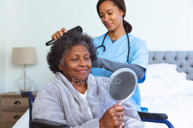 Femme de race mixte âgée passant du temps à la maison, assise dans un fauteuil roulant, tenant un miroir, visitée par une infirmière de race mixte, l'infirmière, l'infirmière se brossant les cheveux — Photo de stock