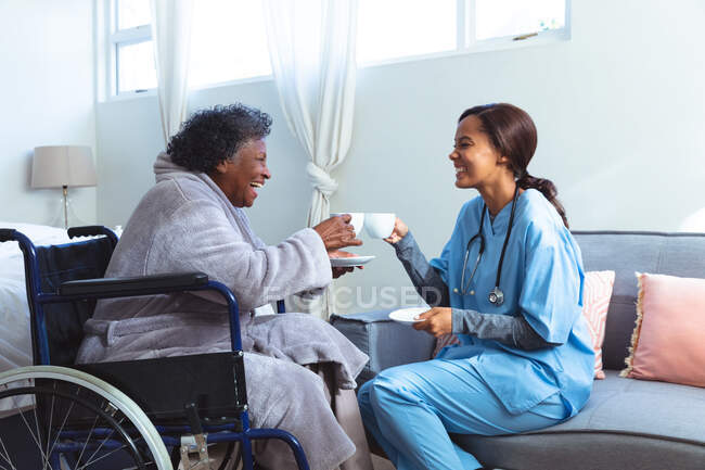Старша змішана жінка проводить час вдома, сидячи на інвалідному візку, відвідуючи змішану расу медсестру, тримаючи чашки і розмовляючи — стокове фото
