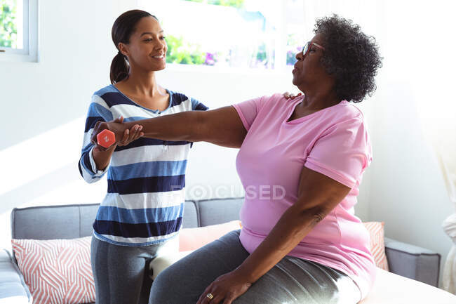Seniorin mit gemischter Rasse verbringt Zeit zu Hause, wird von einer Krankenschwester mit gemischter Rasse besucht, die Frau hält eine Hantel und übt — Stockfoto
