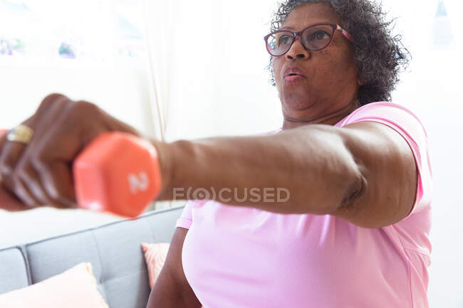 Close up of a senior mixed race woman spending time at home, social distancing and self isolation in quarantine lockdown, holding a dumbbell and exercising — Stock Photo
