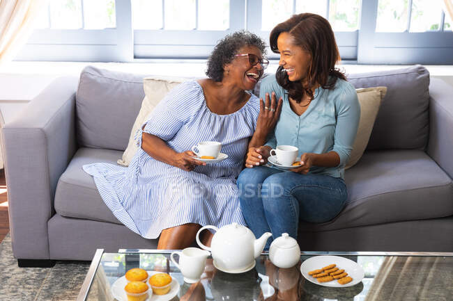 Старша змішана жінка проводить час вдома зі своєю дочкою, соціальне дистанціювання та самоізоляцію в карантинному замку, разом проводячи чай і розмовляючи — стокове фото