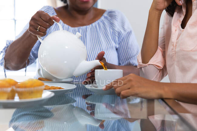 Mitte einer älteren Frau mit gemischter Rasse, die Zeit zu Hause mit ihrer Tochter verbringt, soziale Distanzierung und Selbstisolierung in Quarantäne, zusammen Tee trinkt und redet — Stockfoto