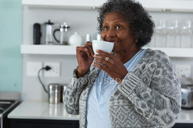 Senior mujer de raza mixta disfrutando de su tiempo en casa, distanciamiento social y auto aislamiento en cuarentena, de pie en su cocina, sosteniendo una taza y sonriendo - foto de stock