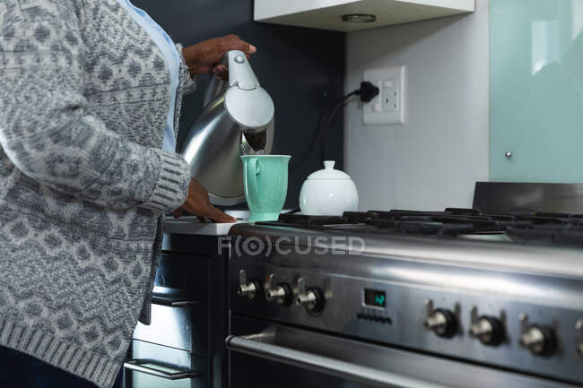 Sección media de la mujer disfrutando de su tiempo en casa, distanciamiento social y auto aislamiento en cuarentena, de pie en su cocina, preparando té - foto de stock