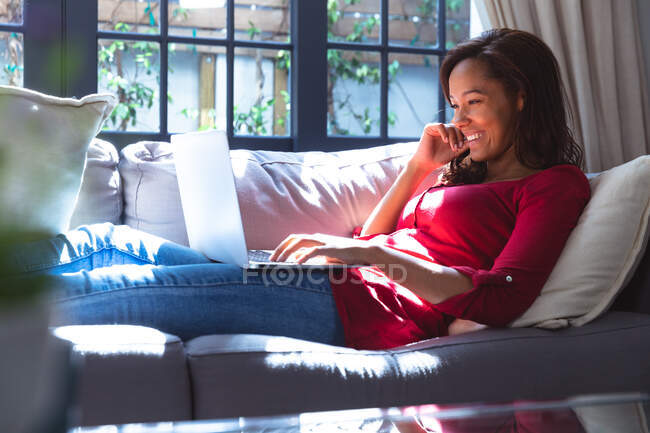 Gemischte Rassenfrau genießt ihre Zeit zu Hause, soziale Distanzierung und Selbstisolierung in Quarantäne, liegt auf einem Sofa, benutzt einen Laptop und lächelt — Stockfoto