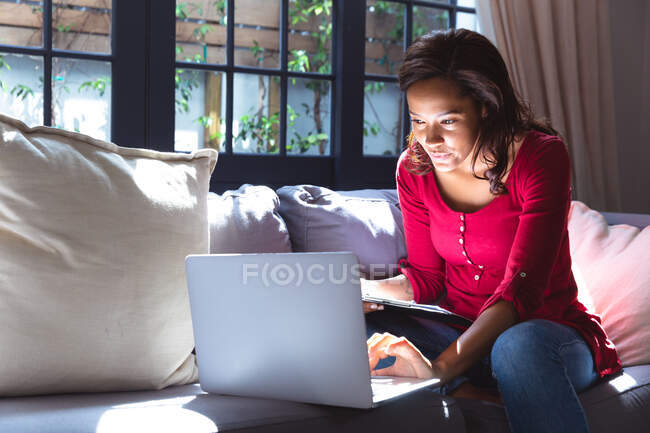 Жінка змішаної раси насолоджується своїм часом вдома, соціальною дистанцією та самоізоляцією в карантинному замку, сидячи на дивані, використовуючи ноутбук — стокове фото