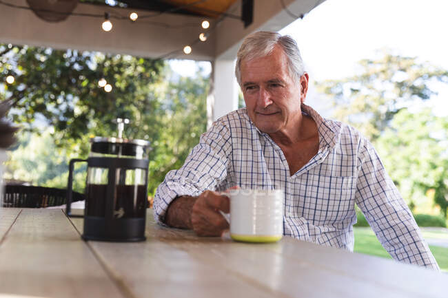 Счастливый пенсионер-европеец дома в саду возле своего дома в солнечный день, сидя за столом с кофеваркой, держа чашку, глядя в сторону и улыбаясь, самоизолируясь во время пандемии коронавируса — стоковое фото