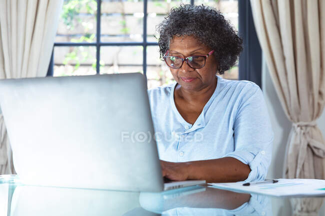 Mujer de raza mixta mayor disfrutando de su tiempo en casa, distanciamiento social y aislamiento en cuarentena, sentada en una mesa, usando una computadora portátil - foto de stock
