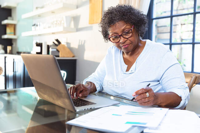Seniorin mit gemischter Rasse genießt ihre Zeit zu Hause, soziale Distanzierung und Selbstisolierung in Quarantäne, sitzt an einem Tisch, benutzt einen Laptop, erledigt Papierkram — Stockfoto