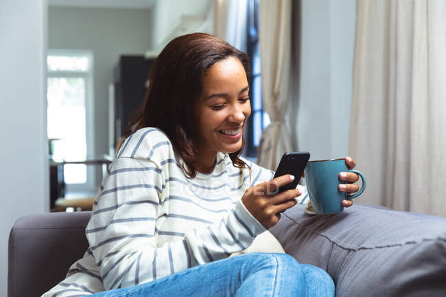 Gemischte Rassenfrau genießt ihre Zeit zu Hause, soziale Distanzierung und Selbstisolierung in Quarantäne, sitzt auf einem Sofa, hält einen Becher in der Hand und benutzt ein Smartphone — Stockfoto