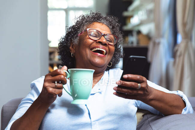 Старша змішана жінка насолоджується своїм часом вдома, соціальною дистанцією та самоізоляцією в карантинному замку, сидячи на дивані, використовуючи смартфон, тримаючи кухоль та посміхаючись — стокове фото