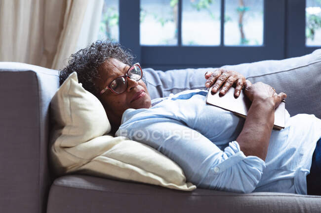 Mulher de raça mista sênior desfrutando de seu tempo em casa, distanciamento social e auto-isolamento em quarentena, deitada em um sofá, dormindo, segurando um livro — Fotografia de Stock