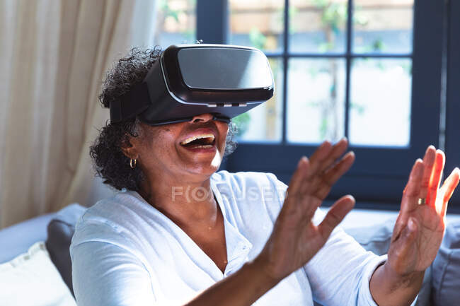 Старша змішана жінка насолоджується своїм часом вдома, соціальною дистанцією та самоізоляцією в карантині, сидячи на дивані, одягаючи окуляри vr та торкаючись екрану віртуальної реальності — стокове фото