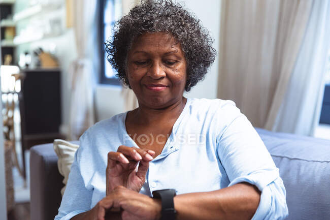 Femme de race mixte âgée appréciant son temps à la maison, la distance sociale et l'isolement personnel en quarantaine verrouillé, assis sur un canapé, en utilisant une montre intelligente — Photo de stock