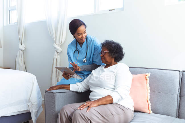 Femme âgée métisse passant du temps à la maison, visitée par une infirmière métisse, utilisant une tablette — Photo de stock
