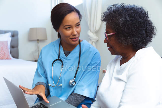 Senior mujer de raza mixta pasar tiempo en casa, ser visitado por una enfermera de raza mixta, utilizando un ordenador portátil y hablando - foto de stock
