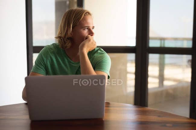 Homem caucasiano sentado ao lado de uma mesa, usando um laptop e olhando para longe. Distanciamento social e auto-isolamento em quarentena. — Fotografia de Stock