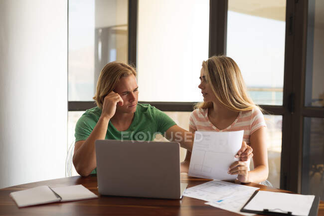 Couple caucasien assis près d'une table, à l'aide d'un ordinateur portable, écrire sur une feuille de papier. Distance sociale et isolement personnel en quarantaine. — Photo de stock