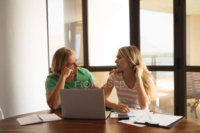Caucasian couple sitting by a table, using a laptop, writing on a sheet of paper and discussing. Social distancing and self isolation in quarantine lockdown. — Stock Photo
