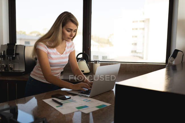 Mujer caucásica de pie junto a una mesa, usando una computadora portátil y escribiendo en una hoja de papel. Distanciamiento social y autoaislamiento en cuarentena. - foto de stock