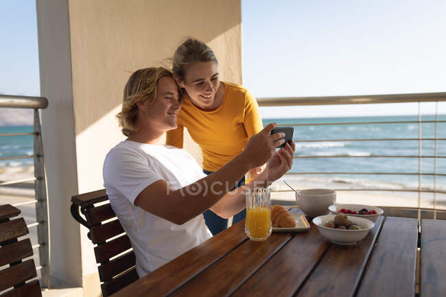 Caucasian couple sitting and standing by a table, a man is showing a woman something on his smartphone. Social distancing and self isolation in quarantine lockdown. — Stock Photo
