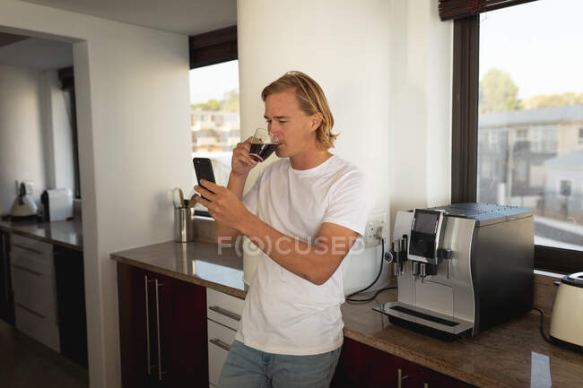 Homme caucasien debout dans une cuisine, boire du café et en utilisant un smartphone. Distance sociale et isolement personnel en quarantaine. — Photo de stock