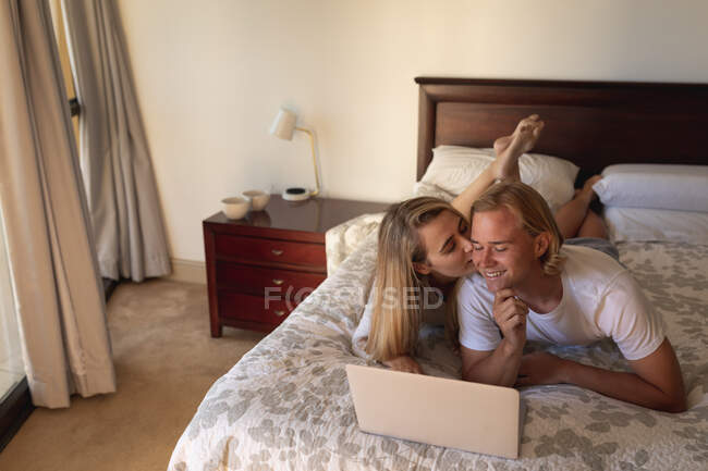 Couple caucasien couché sur le lit ensemble, à l'aide d'un ordinateur portable, une femme embrasse un homme sur sa joue. Distance sociale et isolement personnel en quarantaine. — Photo de stock