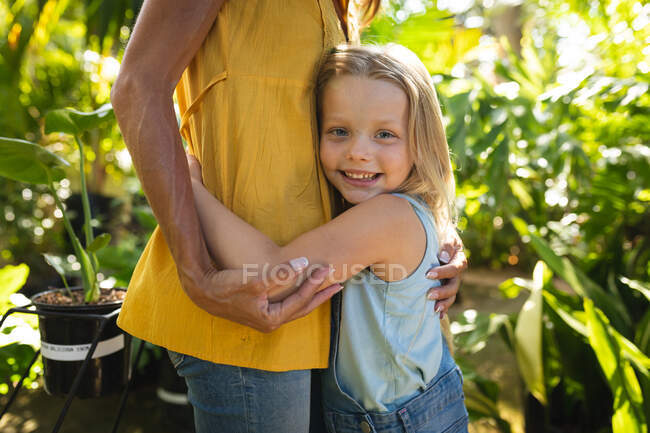 Средний вид на кавказскую женщину и ее дочь, наслаждающиеся временем вместе в солнечном саду, обнимающиеся, дочь смотрит в камеру и улыбается — стоковое фото