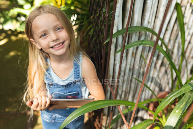 Retrato de una chica caucásica con el pelo largo y rubio disfrutando del tiempo en un jardín soleado, sosteniendo una tableta, mirando a la cámara y sonriendo - foto de stock