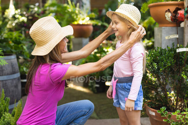 Una mujer caucásica y su hija disfrutando del tiempo juntos en un jardín soleado, mujer arrodillada y poniéndose un sombrero en la cabeza de sus hijas, sonriéndose - foto de stock