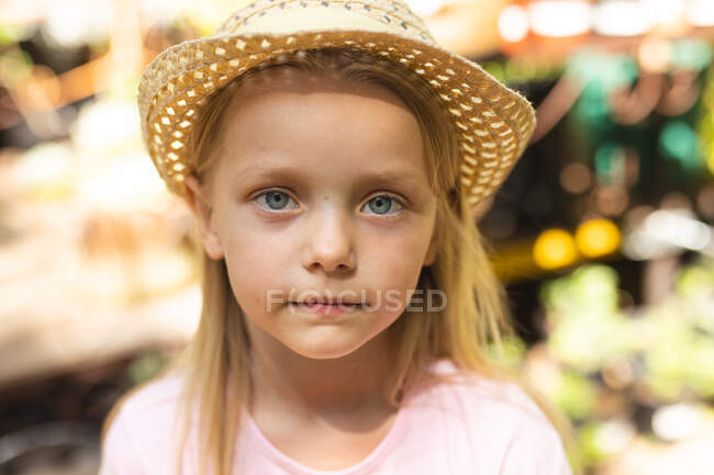 Retrato de una chica caucásica con el pelo largo y rubio, con sombrero de paja, disfrutando del tiempo en un jardín soleado, mirando a la cámara - foto de stock