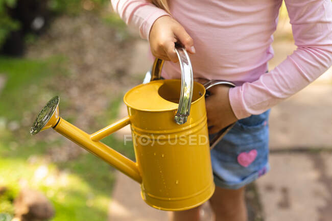 Seção intermediária fechar-se da menina desfrutando do tempo em um jardim ensolarado, explorando, segurando uma lata de rega amarela — Fotografia de Stock