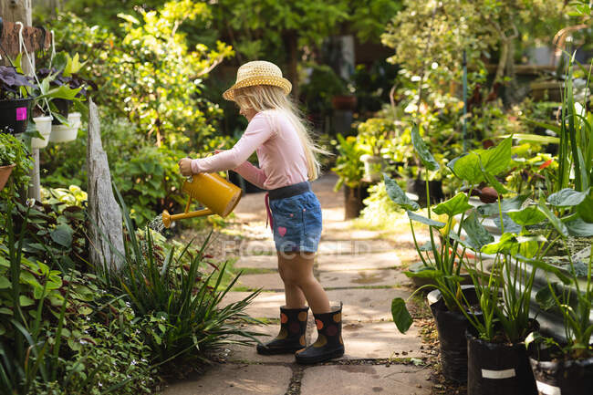 Una chica caucásica con el pelo largo y rubio disfrutando del tiempo en un jardín soleado, explorando, regando plantas con regadera, usando un sombrero de paja - foto de stock