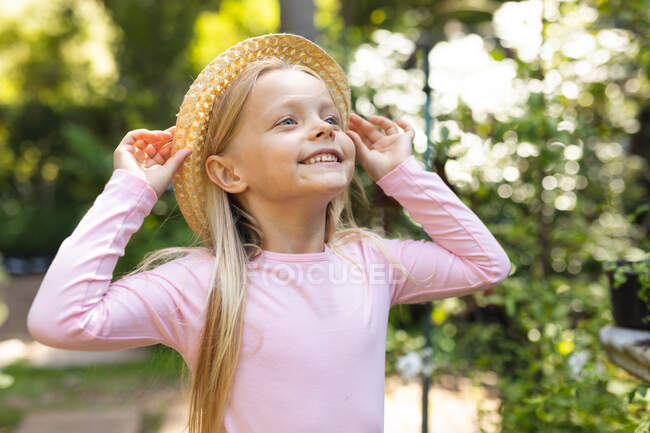 A Caucasian girl with long blonde hair, wearing a straw hat, enjoying time in a sunny garden, looking away and smiling — Stock Photo