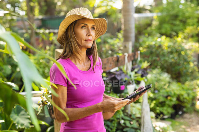 Una mujer caucásica vistiendo una camiseta rosa y un sombrero de paja, disfrutando del tiempo en un jardín soleado, usando una tableta - foto de stock