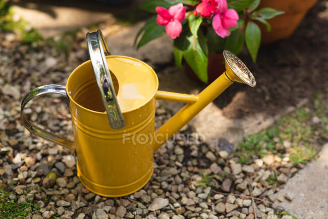 Close up of a yellow watering can used for watering plants placed in a sunny garden next to a pink flower in a pot — Stock Photo