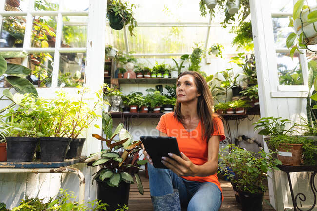 A Caucasian woman with long hair enjoying time in the sunny garden, sitting in the doorway of a greenhouse, using a tablet computer and looking away — Stock Photo