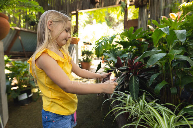 Uma menina caucasiana com longos cabelos loiros desfrutando do tempo em um jardim ensolarado, explorando, tocando folhas de plantas e olhando para elas — Fotografia de Stock