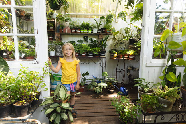 Retrato de uma menina caucasiana com longos cabelos loiros desfrutando do tempo em um jardim ensolarado, de pé na entrada de uma estufa, olhando para a câmera e sorrindo — Fotografia de Stock