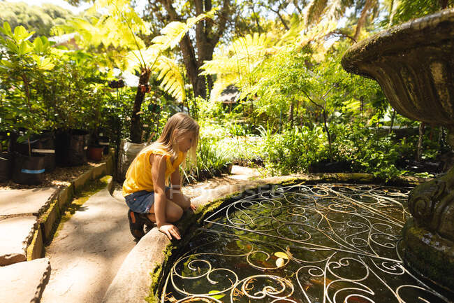 Una chica caucásica con el pelo largo y rubio explorando en un jardín soleado, sentada junto a un estanque y mirando el agua - foto de stock