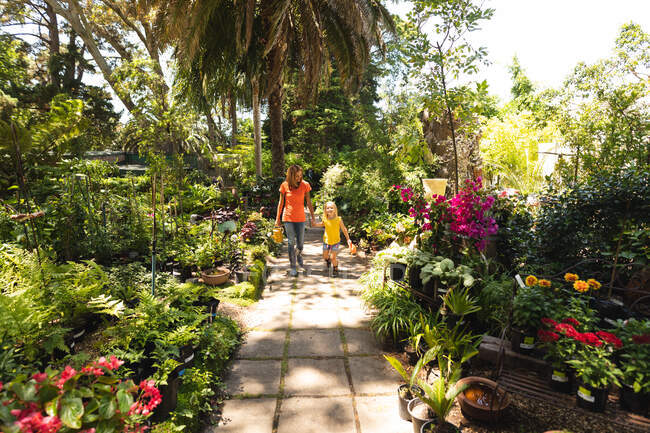 A Caucasian woman and her daughter enjoying time together in a sunny garden, holding hands and walking along a path between plants — Stock Photo