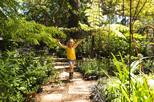 Una chica caucásica con el pelo largo y rubio, disfrutando del tiempo en un jardín soleado, levantando y agitando los brazos corriendo y sonriendo - foto de stock