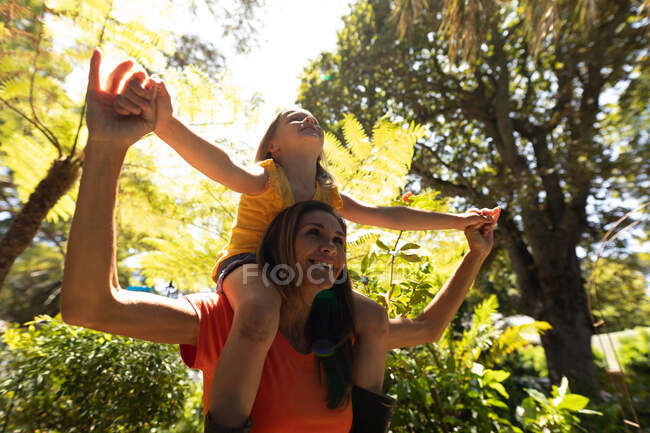 A Caucasian woman and her daughter enjoying time together in a sunny garden, the girl sitting on shoulders of her mother and smiling — Stock Photo