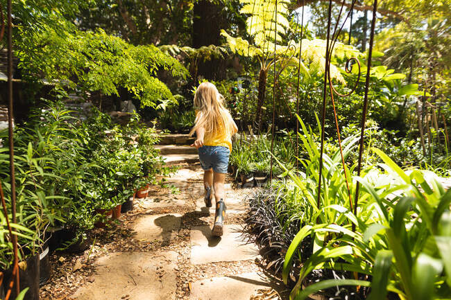 Una chica caucásica con el pelo largo y rubio, disfrutando del tiempo en un jardín soleado, corriendo excitada a lo largo de un camino entre plantas - foto de stock