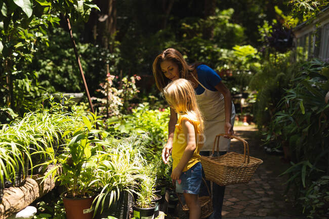 A Caucasian woman wearing an apron and her daughter enjoying time together in a sunny garden, looking at plants together and carrying a selection of plants in baskets — Stock Photo