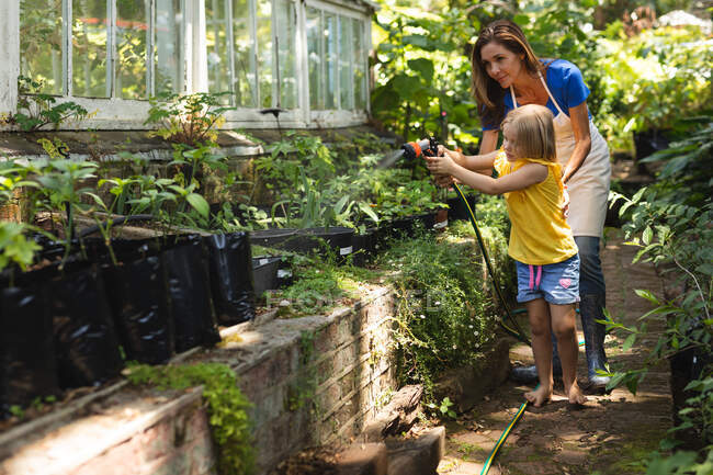 Eine Kaukasierin und ihre Tochter genießen die gemeinsame Zeit in einem sonnigen Garten und gießen die Pflanzen mit einem Gartenschlauch — Stockfoto