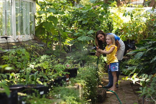 Una mujer caucásica y su hija disfrutando del tiempo juntos en un jardín soleado, usando una manguera de jardín para regar las plantas - foto de stock