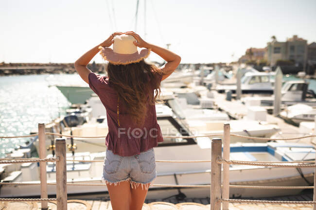 Una adolescente caucásica, con un sombrero de paja, disfrutando de su tiempo en un paseo marítimo, en un día soleado, sosteniendo la cabeza, mirando hacia otro lado - foto de stock