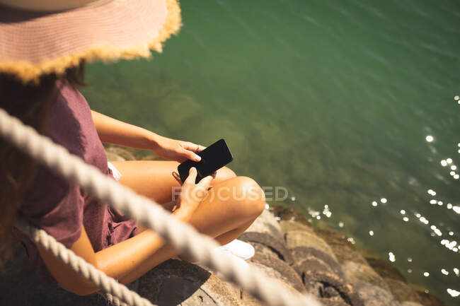 Над плечом кавказская девочка в соломенной шляпе, наслаждающаяся прогулкой, в солнечный день, сидя и пользуясь смартфоном — стоковое фото